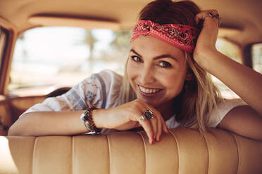 Lächelnde Frau, die sich im Auto entspannt und in die Kamera schaut. Weiblicher Hipster, der sich in einem Oldtimer auf einem Road Trip entspannt. - JLPSF02877