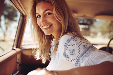 Portrait of smiling woman sitting in front passenger seat of a car and looking back at camera. Woman traveling by a old car, enjoying the ride. - JLPSF02881