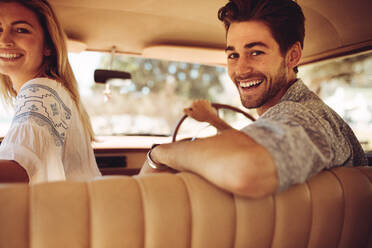 Handsome young man driving the car with his girlfriend sitting by, both looking back and smiling. Couple enjoying on a road trip. - JLPSF02882