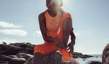 Afrikanische Frau schnürt Laufschuhe im Freien am felsigen Strand. Fitness-Frau macht sich an einem sonnigen Morgen für einen Lauf am Strand bereit. - JLPSF02909
