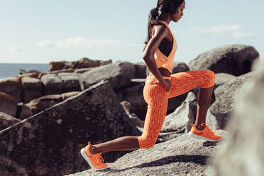 Afrikanische Läuferin, die ihre Beine auf den Felsen ausstreckt. Frau wärmt sich vor dem Freilufttraining am felsigen Strand auf. - JLPSF02912