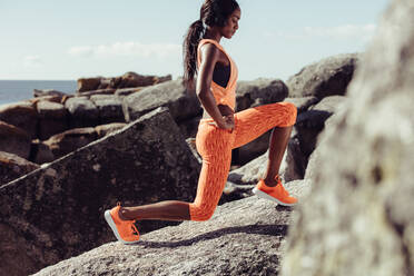 Eine Frau macht Aufwärmübungen auf den Felsen am Strand. Eine Sportlerin dehnt ihren Körper. - JLPSF02914