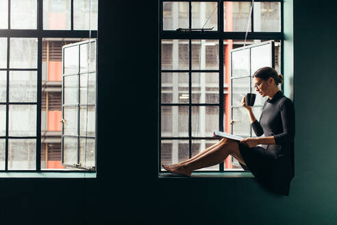 Asiatische Frau sitzt auf der Fensterbank, liest ein Buch und trinkt Kaffee. Frau entspannt sich drinnen mit einer Zeitschrift und trinkt Kaffee. - JLPSF02933