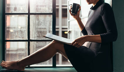 Cropped image of woman with cup of coffee and magazine sitting on window sill. Female drinking coffee and reading fashion magazine. - JLPSF02935