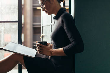 Asiatische Frau mit Kaffee in der Hand und einer Zeitschrift in einem Haus. Junge Frau sitzt mit einem Buch und einer Tasse Kaffee auf der Fensterbank. - JLPSF02936