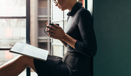 Abgeschnittene Aufnahme einer asiatischen Frau, die mit einer Zeitschrift und einer Tasse Kaffee auf der Fensterbank sitzt. Eine Frau liest ein Buch und trinkt eine Tasse Kaffee, während sie am Fenster sitzt. - JLPSF02937