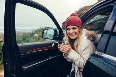 Schöne lächelnde Frau in ihrem Auto, die Kaffee trinkt. Glückliche junge Frau, die eine Tasse Kaffee hält und wegschaut, während sie in ihrem Auto sitzt. - JLPSF02945