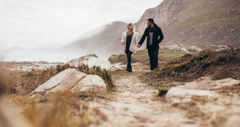 Verliebtes Paar in warmer Winterkleidung, das am Strand spazieren geht. Junger Mann und Frau halten sich an der Hand und spazieren an einem Wintertag am Meer. - JLPSF02955