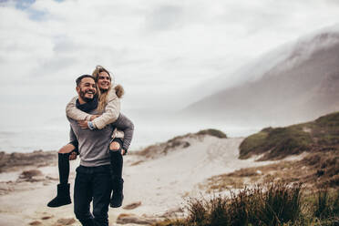 Man carrying girlfriend on his back at winter beach. Young couple enjoying holidays at the seashore. - JLPSF02956