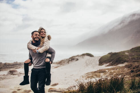 Mann trägt seine Freundin auf dem Rücken am Winterstrand. Junges Paar genießt den Urlaub am Meer. - JLPSF02956
