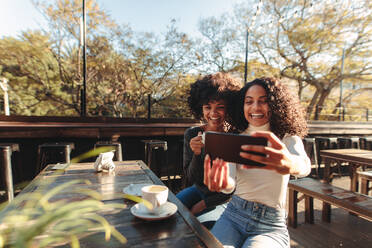 Zwei junge Frauen machen ein Selfie mit einem Handy in einem Café. Freunde sitzen mit Kaffeetassen auf dem Tisch und haben Spaß mit einem Smartphone. - JLPSF02963