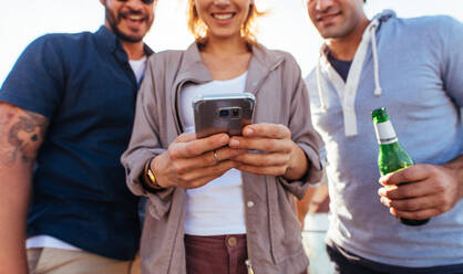 Gruppe von Leuten auf einer Party, die sich ein Video auf dem Handy ansehen. Drei junge Freunde stehen zusammen im Freien und schauen auf ihr Smartphone. - JLPSF02969