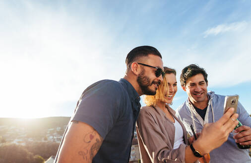 Gruppe von Millennials, die ein Mobiltelefon auf einer Party im Freien benutzen. Lächelnde junge Männer und Frauen, die auf einem Dach stehen und ein Mobiltelefon benutzen. - JLPSF02971