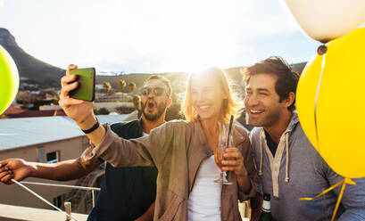 Gruppe von Freunden, die ein Selfie auf einem Handy auf einer Dachparty machen. Junge Männer und Frauen mit Getränken, die ein Selbstporträt auf einem Smartphone machen. Spaß auf einer Dachparty. - JLPSF02973
