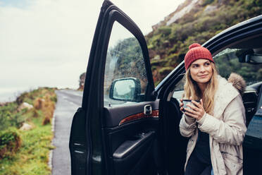 Beautiful female standing by the car with coffee. Smiling woman holding coffee and looking away. - JLPSF02976