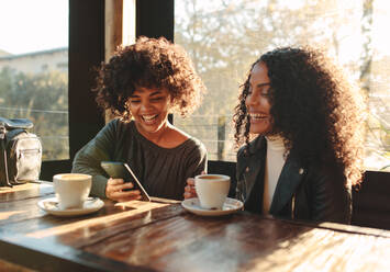 Zwei Frauen lachen und schauen auf ein Handy in einem Café, Freunde sitzen mit Kaffeetassen auf dem Tisch und haben Spaß. - JLPSF02980