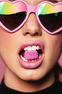 Closeup-Porträt einer jungen Frau mit herzförmiger Sonnenbrille, die ein Bonbon im Mund hält. Schöne rosa Lippen mit einem Stück süßer Süßigkeiten. - JLPSF02981
