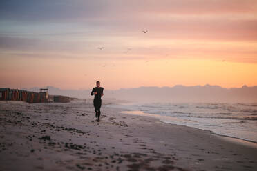 Full length shot of fit male runner running on the beach in morning. Fitness man jogging on the sea shore. - JLPSF03020