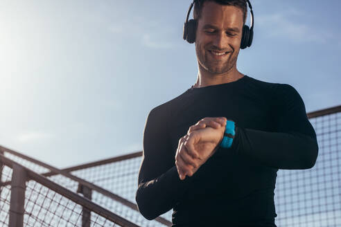 Ein Mann steht im Freien mit Kopfhörern und verwendet eine Smartwatch, um seine Fortschritte zu überwachen. Ein männlicher Sportler ruht sich aus und überprüft seine Leistung auf einer Fitness-Smartwatch. - JLPSF03038
