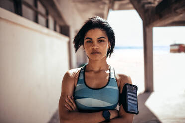 Porträt einer selbstbewussten Sportlerin, die mit verschränkten Armen im Freien steht und in die Kamera blickt. Eine Frau mit einem Mobiltelefon an der Armbinde macht nach dem Lauftraining eine Pause. - JLPSF03043