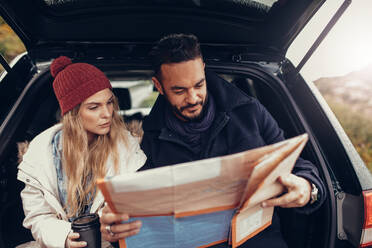 Young couple using a map on a road trip for directions. Young man and woman reading a map while sitting at the back of car. - JLPSF03055