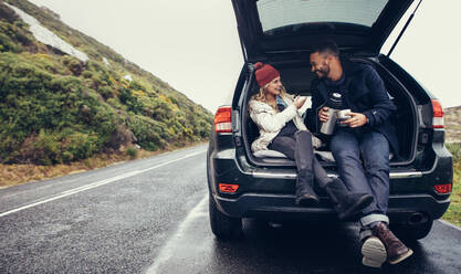 Glückliches junges Paar bei einer Kaffeepause während einer Autofahrt auf dem Lande. Mann und Frau sitzen im Kofferraum und trinken Kaffee. - JLPSF03057