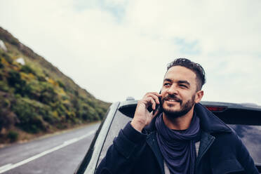 Handsome young man standing outdoors near his car and talking on the mobile phone. Young man on road trip making a phone call. - JLPSF03059