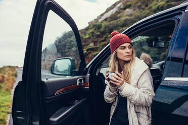 Young woman standing by the car with cup of coffee. Female on road trip holding coffee and looking away. - JLPSF03060
