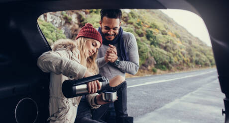 Eine Frau gibt einem Mann, der vor dem Auto steht, heißen Kaffee. Ein Paar auf einer Autoreise macht an einem Wintertag eine Kaffeepause. - JLPSF03068