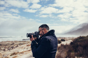 Seitenansicht eines jungen Fotografen, der im Freien fotografiert. Ein Mann, der mit einer Spiegelreflexkamera im Freien fotografiert. Ein Fotograf, der Naturfotografie auf Reisen macht. - JLPSF03076