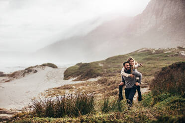 Outdoor shot of man giving woman piggyback on winter beach. Man carrying woman oh his back pointing away. - JLPSF03099