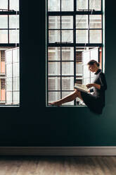 Seitenansicht einer asiatischen Frau, die auf der Fensterbank sitzt und ein Buch liest. Vertikale Aufnahme einer Frau, die an ihrem Bürofenster eine Zeitschrift liest. - JLPSF03101