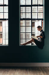 Sideview of asian woman sitting on window sill and reading a book. Vertical shot of female reading a magazine by her office window. - JLPSF03101