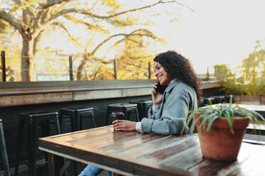 Smiling woman sitting at a table holding a coffee glass and talking over mobile phone. Woman sitting outdoors at a coffee shop. - JLPSF03104