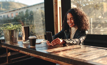 Woman video chatting on mobile phone in a coffee shop. Smiling woman sitting at a table with coffee glass and laptop. - JLPSF03106