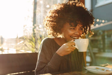 Lächelnde Frau, die eine Kaffeetasse an ihr Gesicht hält und in einem Café sitzt. Lockenköpfige Frau trinkt Kaffee. - JLPSF03118