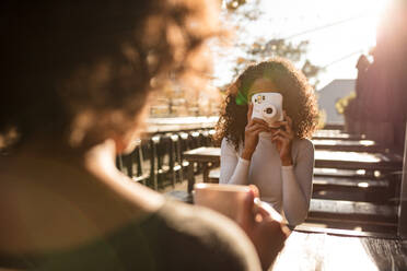 Woman taking photo of her friend using an instant digital camera. Woman holding a coffee cup being photographed by her friend outdoors. - JLPSF03123