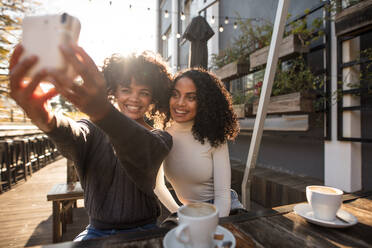 Zwei lächelnde Frauen, die mit einer digitalen Sofortbildkamera in einem Café ein Selfie machen. Freunde sitzen mit Kaffeetassen auf dem Tisch und haben Spaß mit einer Kamera. - JLPSF03125