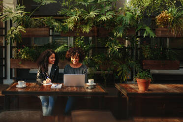 Two women sitting with a laptop and notepads at a coffee table. Business colleagues working while having coffee at a coffee shop. - JLPSF03128