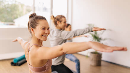 Lächelnde, sportliche Frau, die im Yoga-Kurs die Krieger-Pose einnimmt. Glückliche junge Frau, die im Fitnessstudio Yoga übt. - JLPSF03147
