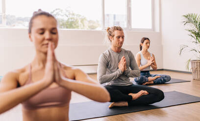 People meditating while sitting in room. Young man practicing yoga in gym class with people sitting around. - JLPSF03151