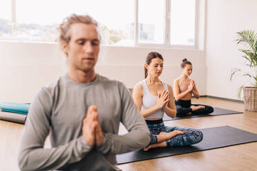 Woman practicing yoga in gym class with people sitting around. People in lotus pose meditating while sitting in room. - JLPSF03153