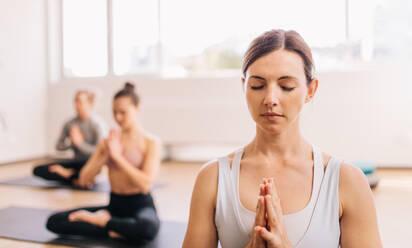 Gesunde Frau macht Yoga im Fitnessstudio mit Menschen im Hintergrund. Menschen meditieren im Fitnessstudio. - JLPSF03154