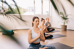 Fitness-Leute sitzen mit gekreuzten Beinen und verschränkten Händen auf dem Boden. Eine Gruppe junger Leute meditiert im Lotussitz beim Yoga-Kurs. - JLPSF03156