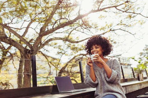 Lockenköpfige Frau sitzt im Freien mit Kaffeetasse in der Hand. Glückliche junge Frau sitzt im Freien mit Laptop und Kaffee. - JLPSF03165