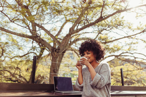 Lockenköpfige Frau sitzt im Freien mit Kaffeetasse in der Hand. Glückliche junge Frau sitzt im Freien mit Laptop und Kaffee. - JLPSF03166