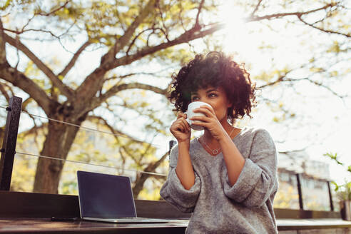 Lockenköpfige Frau sitzt im Freien mit Kaffeetasse in der Hand. Glückliche junge Frau sitzt im Freien mit Laptop und Kaffee. - JLPSF03167