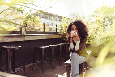 Young woman drinking coffee at a rooftop coffee shop. Smiling curly haired woman enjoying a cup of coffee. - JLPSF03169