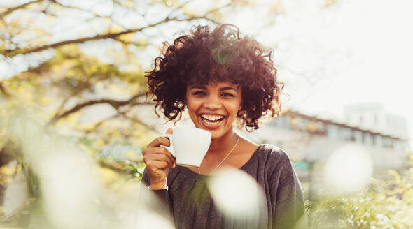 Nahaufnahme einer Frau mit lockigem Haar, die eine Kaffeetasse hält. Fröhliche Frau, die im Freien stehend Kaffee genießt. - JLPSF03174