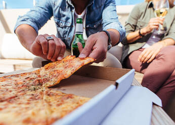 Nahaufnahme einer Pizza auf einem Tisch, auf dem junge Leute sitzen und sich eine Portion abholen. Freunde feiern auf einer Dachterrasse und essen Pizza. - JLPSF03181